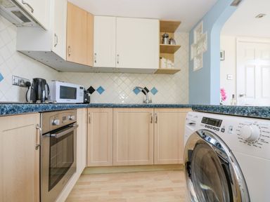 A kitchen with wooden cabinets blue countertops a washing machine oven microwave and kettle at Respryn in Mevagissey