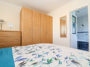 A bedroom with wooden wardrobes a chest of drawers a bed with floral bedding and a view into a tiled bathroom at Respryn in Mevagissey