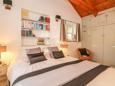 A bedroom with a bed pillows bookshelf window curtains lamps wicker chair and built in wardrobes at Pond Cottage in Stithians