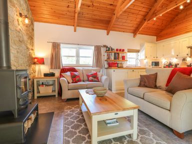 A living room with two sofas, a coffee table, a wood stove, and a kitchen area at Pond Cottage in Stithians