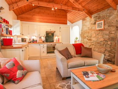 A kitchen area with white cabinets a stone wall beige sofas and a wooden coffee table at Pond Cottage in Stithians