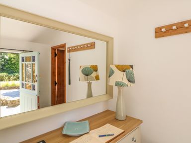 An interior room with a wooden table holding a decorative lamp, a blue dish, and an open notebook under a large wall mirror reflecting an open door at Pond Cottage in Stithians