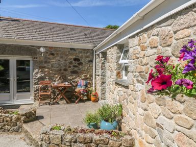 A stone patio with wooden chairs and table with cups and a blanket and potted plants at Pond Cottage in Stithians