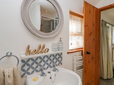 A bathroom sink with two taps a round mirror and a wooden hello sign on a shelf above at Pond Cottage in Stithians