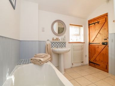 A bathroom with a bathtub towels a pedestal sink a round mirror wooden door and a window at Pond Cottage in Stithians