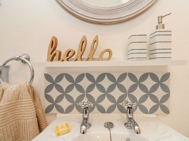 A bathroom sink with two chrome taps a beige towel on a ring a soap bar a wooden hello sign and soap dispensers at Pond Cottage in Stithians