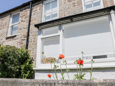An exterior view of a stone cottage with white framed windows and red flowers in front at Calac Cottage in Mousehole