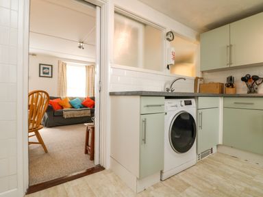 A kitchen with a washing machine green cabinets and utensils next to a living room with a wooden chair and a sofa with colorful cushions at Calac Cottage in Mousehole