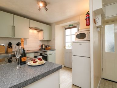 A kitchen with a countertop holding a bottle of sparkling wine glass two plates of scones a refrigerator with a microwave on top and a door with a fire exit sign at Calac Cottage in Mousehole