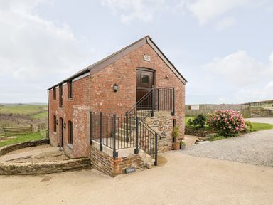 A brick building with stairs and a black metal railing outside surrounded by greenery at Castle Dore Barn in Fowey