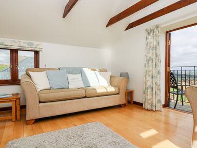 A living room with a beige sofa and cushions next to a window and an open door leading to a balcony at Castle Dore Barn in Fowey