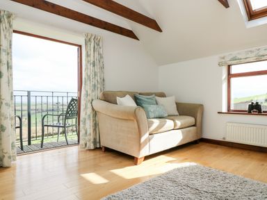A living room with a beige sofa with cushions next to a door opening to a balcony with metal chairs at Castle Dore Barn in Fowey