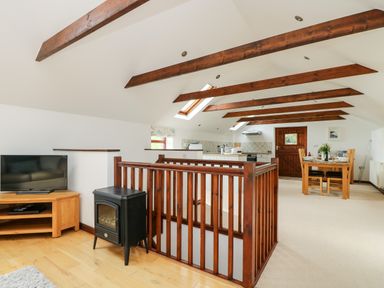 An open-plan room with exposed wooden beams a staircase wooden dining table a television on a wooden stand and a small stove at Castle Dore Barn in Fowey