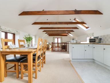A dining area with wooden table and chairs next to a kitchen with white cabinets and beams on the ceiling at Castle Dore Barn in Fowey