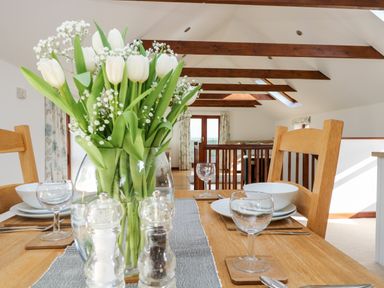 A dining table set with plates bowls glasses and salt and pepper shakers with white flowers in a vase in a room with wooden beams and doors at Castle Dore Barn in Fowey