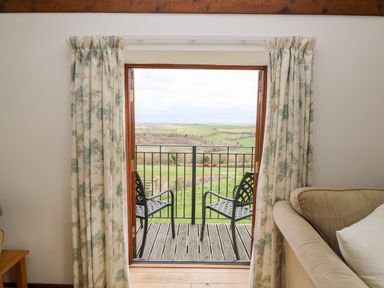 A balcony with two chairs and a table overlooking green fields seen through open doors with floral curtains at Castle Dore Barn in Fowey