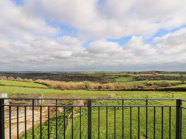 A view of green fields and hills with grazing horses seen from behind a black metal railing at Castle Dore Barn in Fowey