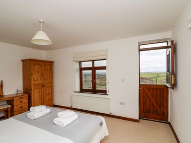 A bedroom with a bed, wooden wardrobe, desk, window, and half open wooden door showing a countryside view at Castle Dore Barn in Fowey