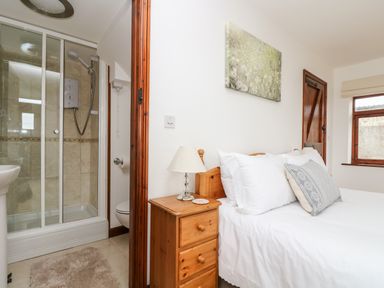 A bedroom with a wooden bed and white bedding next to a bedside table with a lamp and an ensuite shower at Castle Dore Barn in Fowey