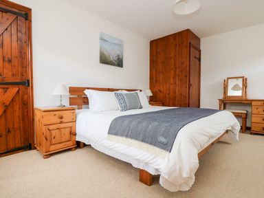 A bedroom with a wooden bed frame white bedding blue throw pillows two bedside tables with lamps a wooden door wooden wardrobes and a dressing table with a mirror at Castle Dore Barn in Fowey
