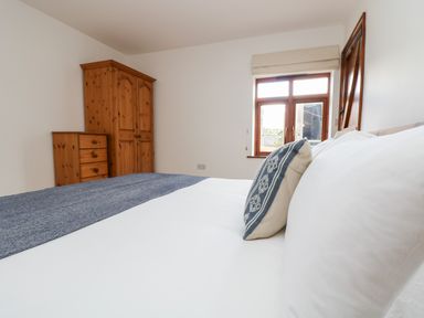 A bedroom with a bed featuring white and blue bedding a wooden wardrobe a wooden chest of drawers and a window at Castle Dore Barn in Fowey
