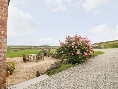 An outdoor patio area with wooden table and chairs next to a gravel pathway and flowering bushes at Castle Dore Barn in Fowey