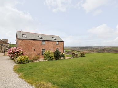 A red brick barn with slate roof surrounded by grass and flowering bushes with a countryside view at Castle Dore Barn in Fowey