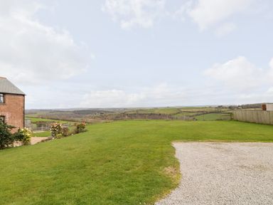 A grassy yard with a gravel area next to a brick house overlooking fields and hills at Castle Dore Barn in Fowey
