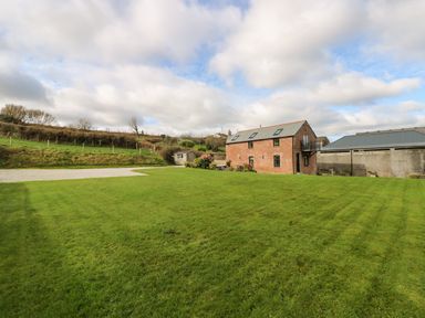 A large lawn with a two-story brick building and a shed in the background at Castle Dore Barn in Fowey