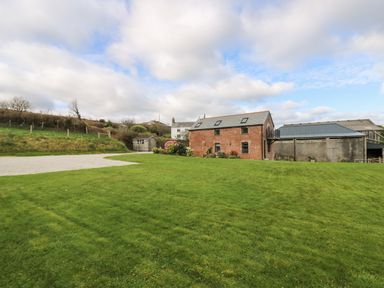 A brick barn with skylights and adjacent buildings surrounded by a lawn and gravel driveway at Castle Dore Barn in Fowey