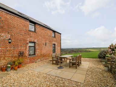 An outdoor patio with a wooden table and six chairs beside a brick building overlooking a green field at Castle Dore Barn in Fowey