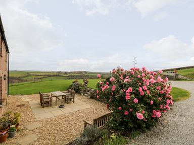 An outdoor patio area with wooden table and chairs surrounded by gravel and flowering bushes at Castle Dore Barn in Fowey