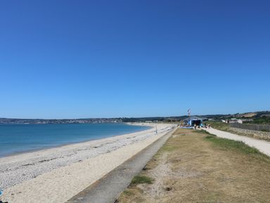 A beach with sand and pebbles next to a path and a cafe building by the shore at Surfside in Marazion