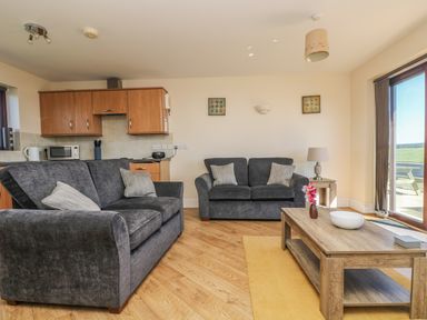 A living room with two grey sofas a wooden coffee table with flowers and a bowl and a kitchen area at Surfside in Marazion