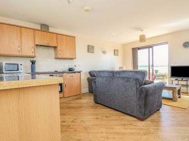 An open plan kitchen and living room with wooden cabinets gray sofas a TV and a sliding glass door at Surfside in Marazion