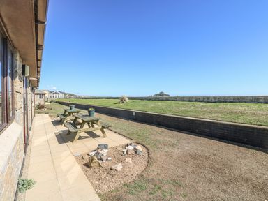 An outdoor patio area with picnic tables and flower pots next to a stone building with a grass embankment in the background at Surfside in Marazion