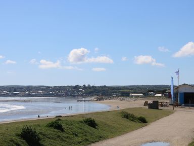 A beach with a sandy shoreline and grassy area with a small building and flags near the water at Surfside in Marazion