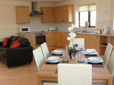 A dining table with plates and glasses in a kitchen with wooden cabinets and a black sofa with red cushions at Sea Shore in Marazion