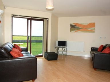 A living room with two black sofas and red cushions a slipcovered ottoman a TV on a stand a glass door leading outside and an abstract painting on the wall at Sea Shore in Marazion