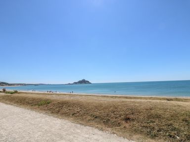 A sandy path with dry grass leading to a beach with people near the shoreline and an island with a castle in the background at Sea Shore in Marazion