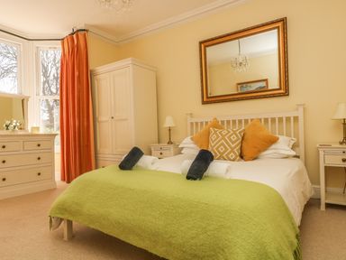 A bedroom with a bed with pillows and towels a green blanket bedside tables lamps a wardrobe a dresser with a mirror and orange curtains at Boskednan Wartha in Penzance
