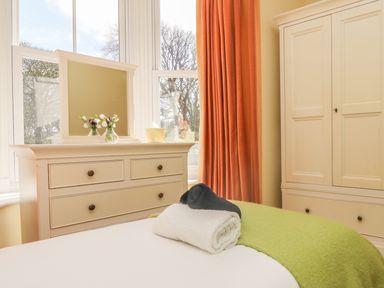 A bedroom with towels on a bed a dresser with mirror and flowers and a wardrobe with orange curtains at Boskednan Wartha in Penzance