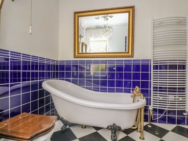 A bathroom with a white clawfoot bathtub blue tiled walls a wooden toilet seat a gold framed mirror and a white towel radiator at Boskednan Wartha in Penzance