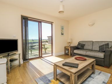 A living room with a sofa a wooden coffee table a television on a glass stand and sliding glass doors leading outside at Seashells in Marazion