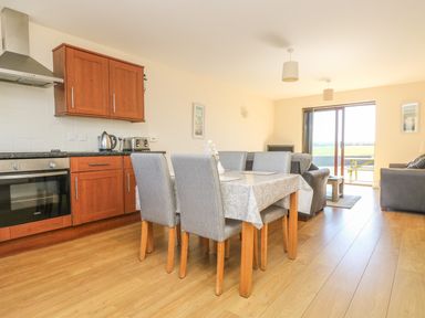 A kitchen and dining area with wooden cabinets a dining table with gray chairs and a living area with sofas and sliding glass door at Seashells Marazion