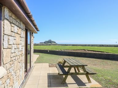 A stone building with a glass door next to a patio with a wooden picnic table and grassy area with a distant hill at Seashells in Marazion