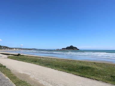 A coastal scene with a sandy path next to grass and a beach with waves and an island with a castle in the background at Seashells in Marazion