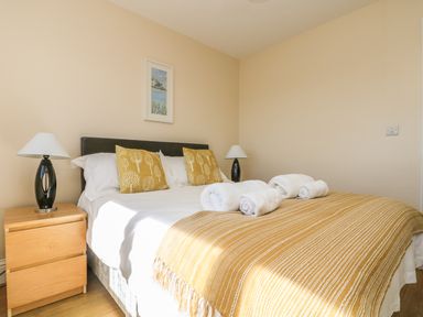 A bedroom with a double bed with yellow cushions and blanket two bedside tables with black lamps and framed artwork on the wall at Seashells in Marazion