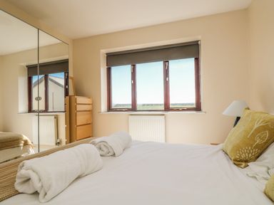 A bedroom with a bed with rolled white towels and yellow pillows near a window with a gray roller blind and a mirrored wardrobe at Seashells in Marazion