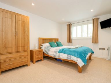 A bedroom with a wooden bed frame wardrobe and nightstand a window with curtains and a wall-mounted television at Rock Pool in Downderry